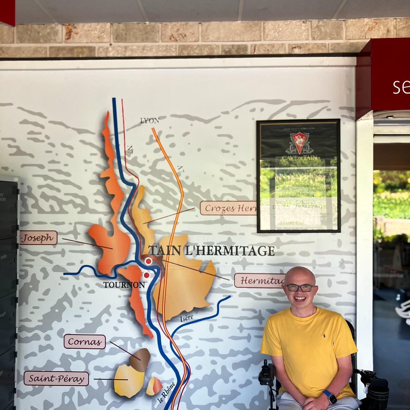 Ryleigh Newman seated beside a large wall map of the Tain-l'Hermitage and Rhone valley wine appellations at the Cave de Tain.