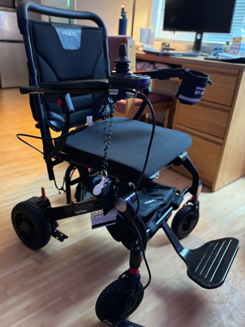 A folding Pride power wheelchair with a black seat, a joystick controller, and a carbon fiber footplate, parked on a wood floor in front of a desk and monitor.