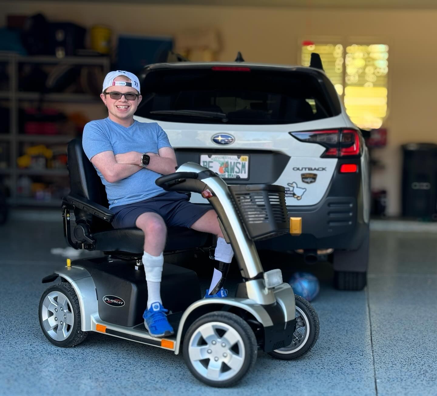 Ryleigh Newman in sunglasses and a cap, arms crossed and smiling, seated on a four-wheel mobility scooter in a garage with an ankle brace on one leg, parked behind a Subaru Outback.